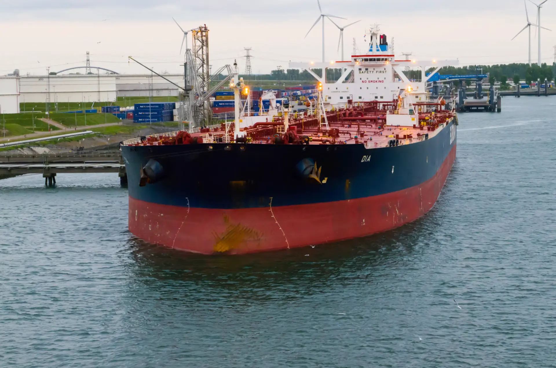 Oil tanker navigating an industrial harbour with storage tanks and cranes